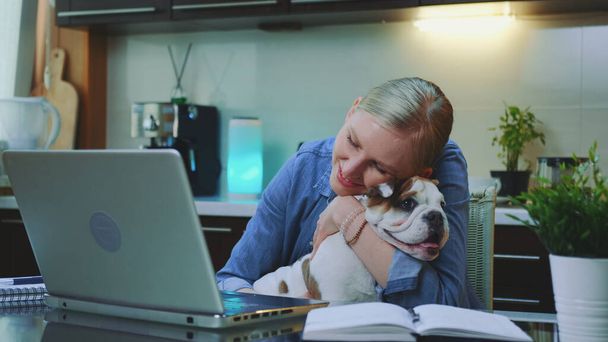 Lächelnde Frau streichelt und umarmt die Bulldogge, während sie am Computer sitzt - Foto, Bild