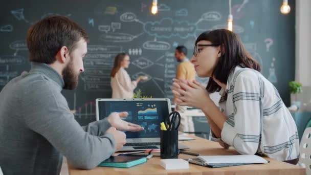 Man and woman discussing work looking at laptop screen while coworkers witing on blackboard - Filmagem, Vídeo