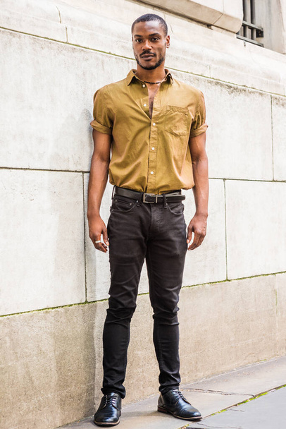 ニューヨークの若いアフリカ系アメリカ人男性の大学生の肖像、ひげ、緑の半袖シャツ、黒パンツ、革の靴を着て、壁に対してオフィスビルの外に立って - 写真・画像