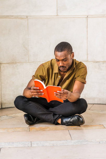Young African American Man with beard studying in New York City, wearing green short sleeve shirt, black pants, leather shoes, crossing legs, sitting on marble ground on campus, reading red book - Foto, imagen