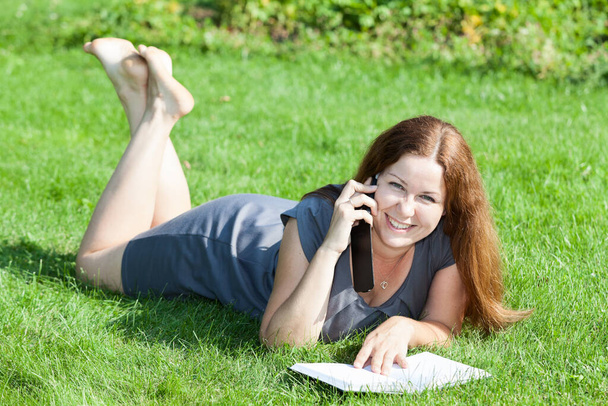 Portrait of red hair Caucaian woman lying on green grass lawn with a book, calling on smartphone - Фото, изображение