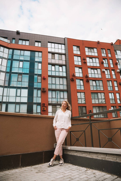 Full length portrait of stylish pretty cheerful blonde woman posing on the street near business center buildings, wearing dress coat and sneakers, fashion lifestyle, positive mood. - Foto, Imagem
