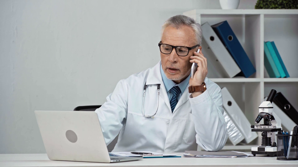 middle aged doctor writing prescription and talking on smartphone  - Footage, Video