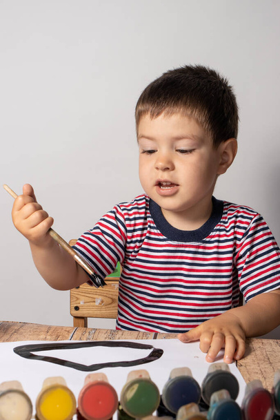 Egy 2-3 éves kisfiú csíkos, többszínű pólóban guasya festéket rajzol papírra. Szabadidő és szórakozás gyerekeknek, gyermekfejlesztés, korai fejlesztés. A kisgyermekek színes rajzai - Fotó, kép