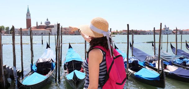 girl with straw hat and fuchsia backpack in Venice looks at the island of San Giorgiowith basilica and bell tower in the Venetian lagoon - Photo, Image