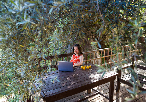Nainen punaisessa mekossa työskentelee puutalon terassilla, Tyttö tutkii kannettavaa tietokonetta, etäopetus, Freelancer työskentelee talon verannalla, etätyö karanteenin aikana. - Valokuva, kuva