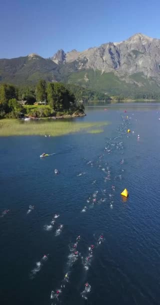 Vzdušný pohled na plavce v triatlonové soutěži Ironman přes jezero Nahuel Huapi za slunečného letního dne. Obklopen horami, vegetací a čluny. - Záběry, video