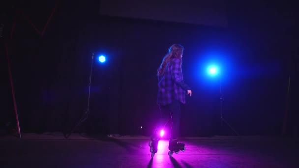 Pretty young woman professionally skates while spinning in night studio on rear and front wheel under blue and pink spotlight. Sports girl is casting on stage and shows the technique of roller skating - Footage, Video