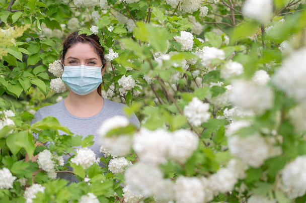 An unhappy red-haired woman in a medical mask stands next to a blossoming apple tree in a park. Seasonal allergic rhinitis. The girl suffers from an allergy to pollen. - Фото, изображение