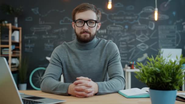 Portrait of young man in glasses looking at camera sitting at desk, laptop computer is visible - Materiał filmowy, wideo