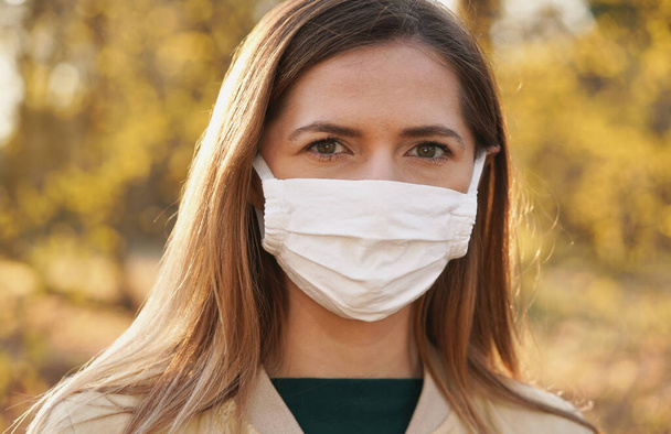 Young woman wearing white cotton virus mouth nose mask in park, sun lit blurred yellow leaves trees, in background, closeup face portrait - Photo, Image