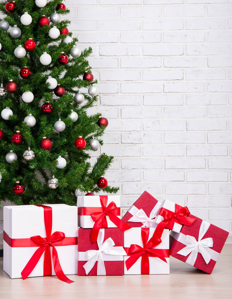 Caixas de presente sob árvore de natal decorada com bolas coloridas sobre parede de tijolo branco
 - Foto, Imagem