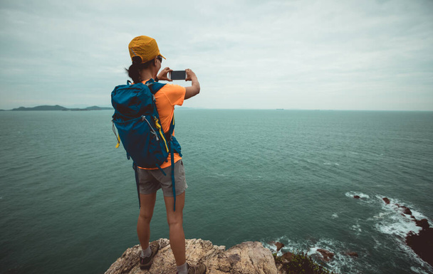 Woman hiker taking photo with smartphone on seaside cliff edge - 写真・画像
