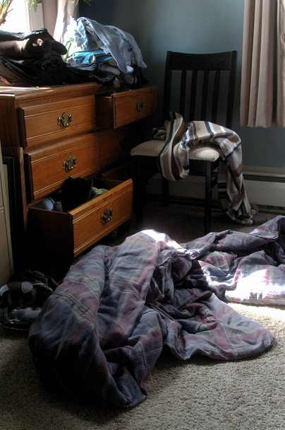 Cajones abiertos de una cómoda, manta desplegada en el suelo y desorden en la esquina de un dormitorio iluminado por el sol - Foto, imagen
