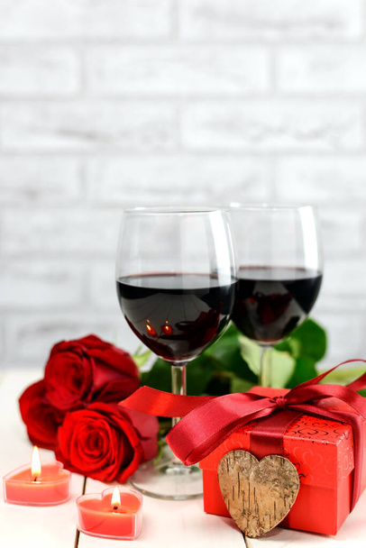Valentinstag-Konzept. Zwei Gläser Wein, rote Rosen, Geschenkbox, Holzherz und brennende Kerzen auf einem weißen Holztisch. Selektiver Fokus. - Foto, Bild