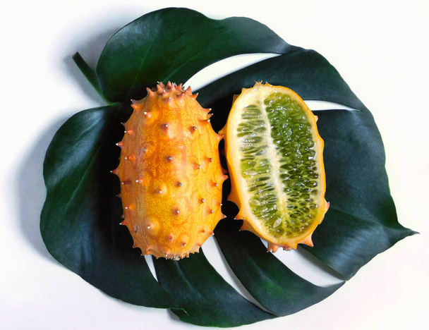 Egy egész és fél kiwano gyümölcs középpontjában egy zöld digitalia monstera levél és világít a természetes napfény - Fotó, kép