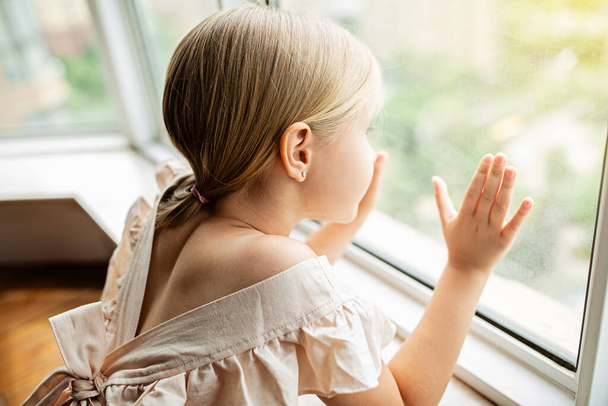Stylowa mała dziewczynka z blond włosami siedzi w domu w pobliżu okna podczas coronavirus covid-19 samoizolacji - Zdjęcie, obraz