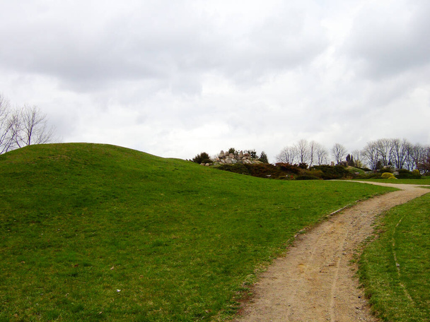 On the left is a green grassy hill and near it a dirt track against a gray sky with clouds. In the background are shit - Foto, immagini