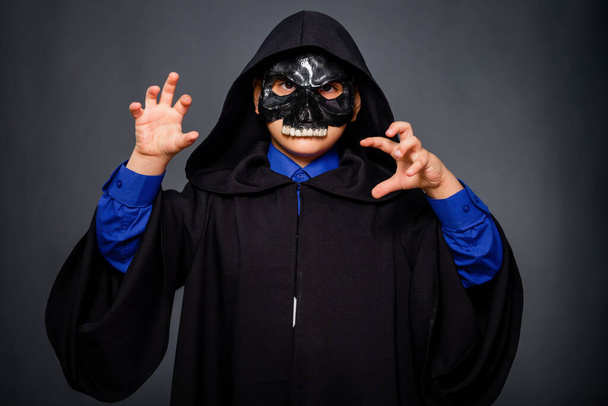 A boy dressed in a black cloak with a hood and a black skull mask, standing on a gray background. A child in a Halloween costume makes threatening gestures, scares. - Fotoğraf, Görsel