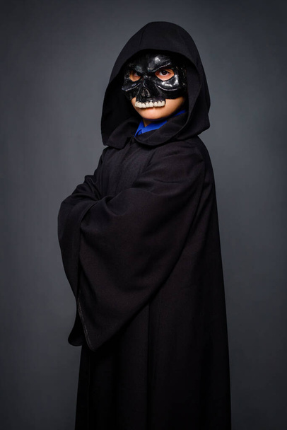 Een jongen in een Halloween kostuum, gekleed in een zwarte mantel met een kap en een zwarte schedel masker, staande op een grijze achtergrond. - Foto, afbeelding