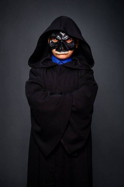Een jongen in een Halloween kostuum, gekleed in een zwarte mantel met een kap en een zwarte schedel masker, staande op een grijze achtergrond. - Foto, afbeelding