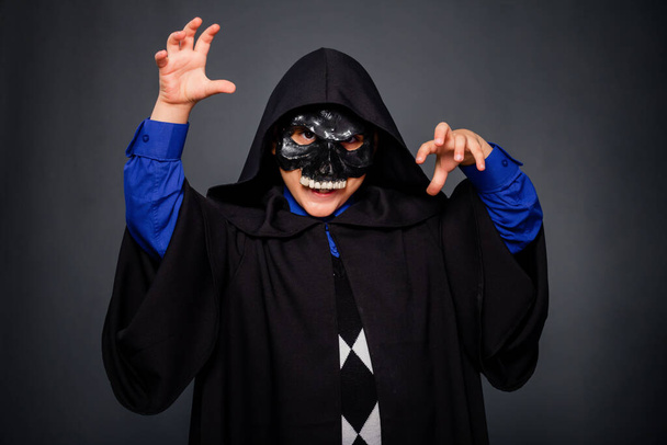 A boy dressed in a black cloak with a hood and a black skull mask, standing on a gray background. A child in a Halloween costume makes threatening gestures, scares. - Photo, image