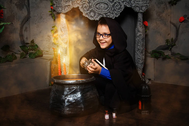 Мальчик-волшебник в очках с волшебной палочкой в руках и в черной мантии с капюшоном готовит зелье в черном котле, сидя в тумане и дыме
. - Фото, изображение
