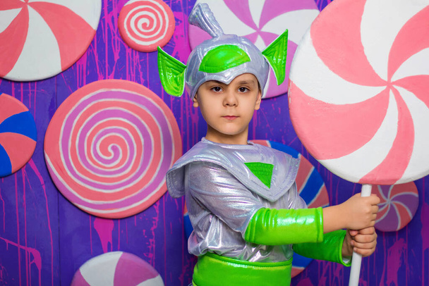 宇宙人の衣装を着た少年がお菓子の壁を背景に大きな飴を手に持っている. - 写真・画像