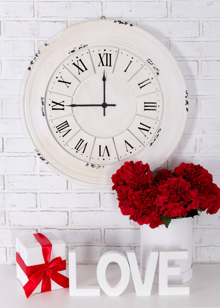 concepto de amor y día de San Valentín - letras de madera que forman la palabra AMOR, flores, regalo y reloj vintage sobre fondo de pared de ladrillo blanco
 - Foto, imagen