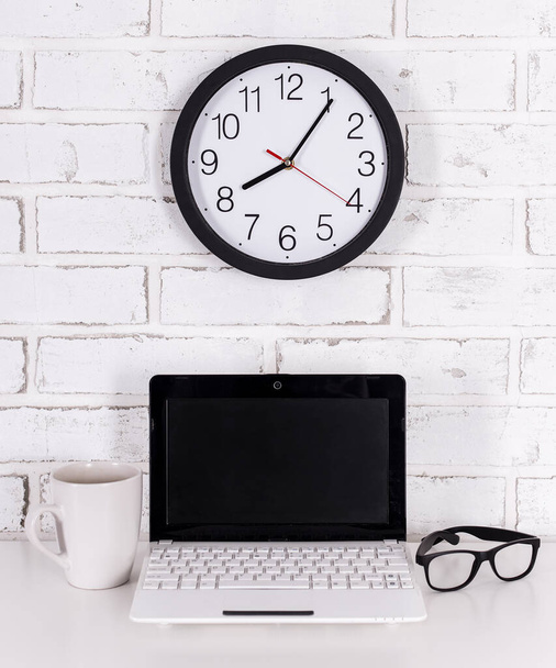 lieu de travail à la maison ou au bureau ordinateur portable moderne, lunettes, tasse de café et horloge de bureau au-dessus du mur de briques - Photo, image