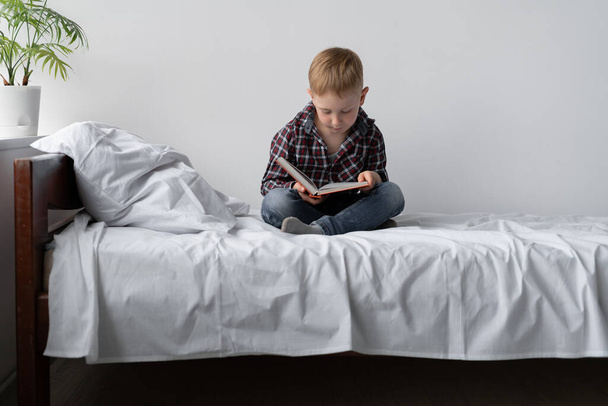 A boy on self-isolation reads a book and stays at home. He is sitting on the bed and his legs are folded. Self study lessons. Rest student from school. Free time and education. Place for text. - Photo, Image