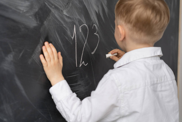 Egy fiú háttal áll egy tábla előtt az iskolában. Matek óra az általános iskolában. Egy iskolás a táblánál krétával írja a számokat. A gyerek keze egy táblán fekszik. Általános iskolások oktatása. A szöveg helye. - Fotó, kép