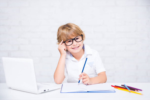 portrét šťastného malého školáka v brýlích, jak dělá domácí úkoly a o něčem přemýšlí - Fotografie, Obrázek