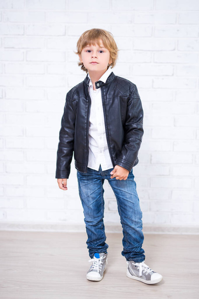 full length portrait of little boy in leather jacket posing over white brick wall background - Fotoğraf, Görsel