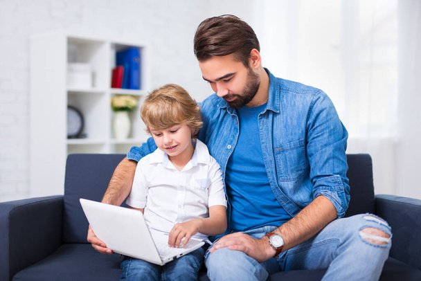 počítač a internet koncept - mladý otec a syn sedí na pohovce s moderním notebookem - Fotografie, Obrázek