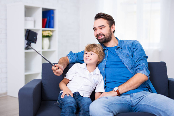 family concept - ο νεαρός πατέρας και ο μικρός γιος του βγάζουν selfie φωτογραφία στο σπίτι - Φωτογραφία, εικόνα