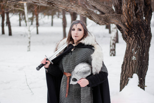 Portrait of a girl of a Viking-era warrior in a chain mail coat and fur with a sword and helmet in her hands. Against the backdrop of the winter forest. - 写真・画像