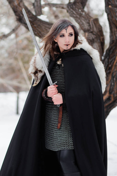  Portrait of a medieval woman, warrior of the Viking Age in the winter forest on the background of a large tree. Dressed in chain mail, cloak, fur, leather pants and boots with a sword in their hands. Battle paint on the face. - Foto, Imagen
