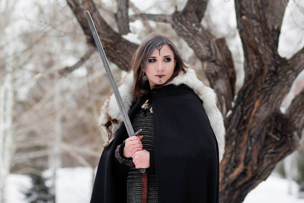  Portret średniowiecznej kobiety, wojowniczki epoki wikingów w zimowym lesie na tle dużego drzewa. Ubrani w kolczugę, pelerynę, futro, skórzane spodnie i buty z mieczem w rękach. Farba bojowa na twarzy. - Zdjęcie, obraz