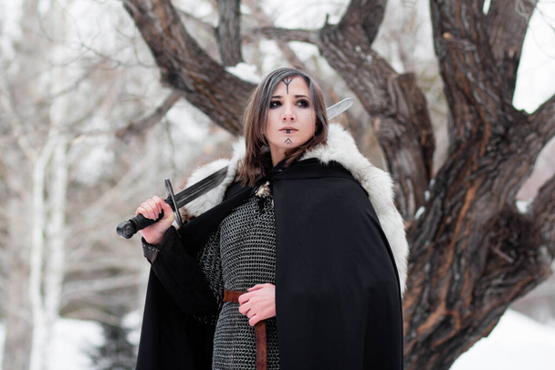  Portré egy középkori nőről, a Viking kor harcosáról a téli erdőben, egy nagy fa hátterében. Láncba verve, köpenyben, bundában, bőrnadrágban és csizmában, karddal a kezükben. Harci festék az arcon.. - Fotó, kép