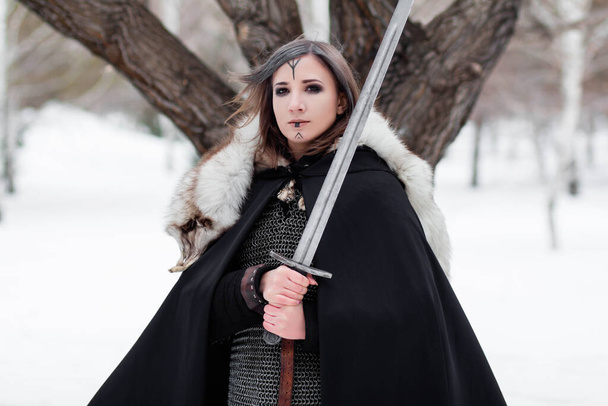  Portrait of a medieval woman, warrior of the Viking Age in the winter forest on the background of a large tree. Dressed in chain mail, cloak, fur, leather pants and boots with a sword in their hands. Battle paint on the face. - 写真・画像