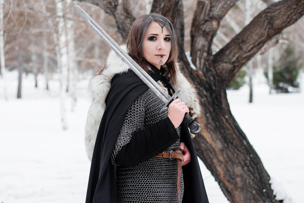  Portrait of a medieval woman, warrior of the Viking Age in the winter forest on the background of a large tree. Dressed in chain mail, cloak, fur, leather pants and boots with a sword in their hands. Battle paint on the face. - Fotoğraf, Görsel