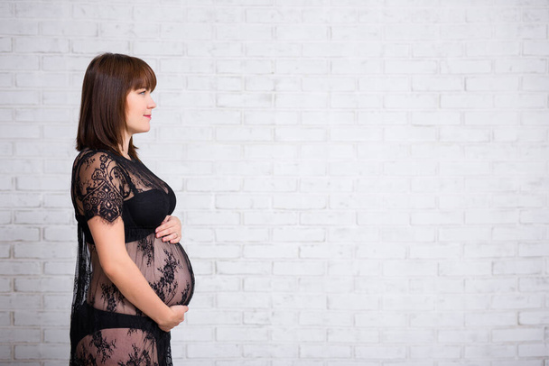 πλαϊνή άποψη της εγκύου γυναίκας σε μαύρο φόρεμα δαντέλα ποζάρουν πάνω από λευκό φόντο τούβλο τοίχο με αντίγραφο χώρο - Φωτογραφία, εικόνα