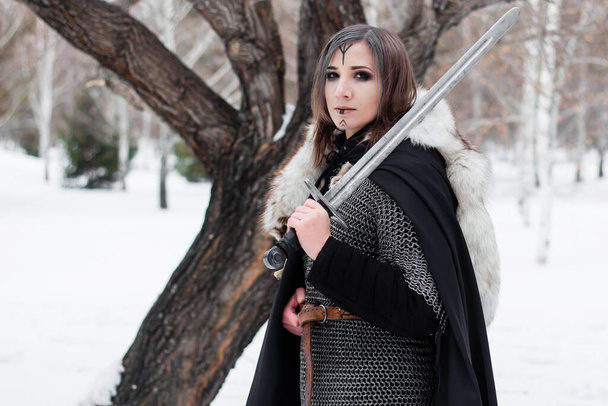  Portrait of a medieval woman, warrior of the Viking Age in the winter forest on the background of a large tree. Dressed in chain mail, cloak, fur, leather pants and boots with a sword in their hands. Battle paint on the face. - Foto, imagen