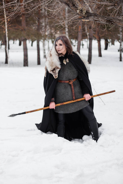 The woman is a medieval warrior of the Viking Age in the winter in the forest. Dressed in chain mail, cloak, fur,  leather pants and boots with a spear in their hands. Battle coloring on the face. - Foto, imagen