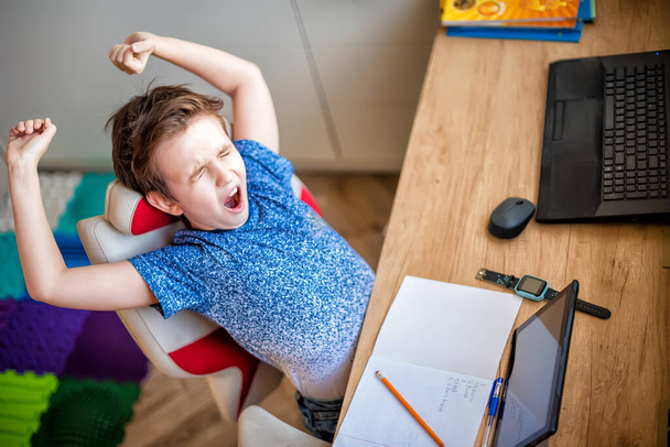 Мальчик зевает Уик, занимаясь дома в детской комнате. На столе есть блокнот, карандаш, ноутбук, планшет и умные часы
. - Фото, изображение