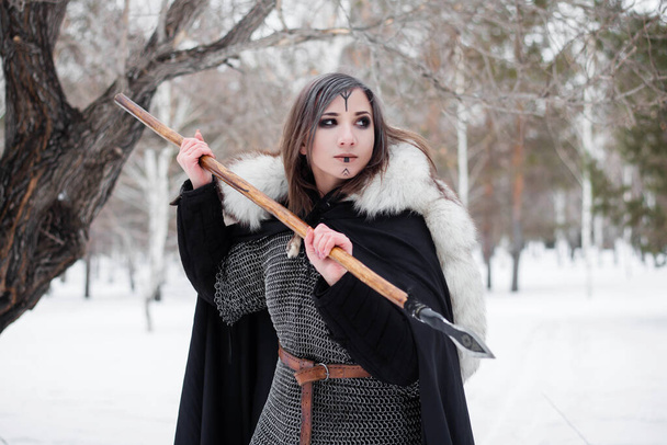 Kobieta jest średniowiecznym wojownikiem epoki wikingów zimą w lesie. Ubrani w kolczugę, pelerynę, futro, skórzane spodnie i buty z włócznią w rękach. Bitwa kolorowanki na twarzy. - Zdjęcie, obraz