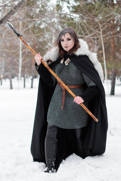 The woman is a medieval warrior of the Viking Age in the winter in the forest. Dressed in chain mail, cloak, fur,  leather pants and boots with a spear in their hands. Battle coloring on the face. - Φωτογραφία, εικόνα