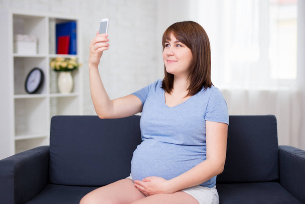 szczęśliwy młody kobieta w ciąży biorąc selfie zdjęcie ze smartfonem w salonie - Zdjęcie, obraz