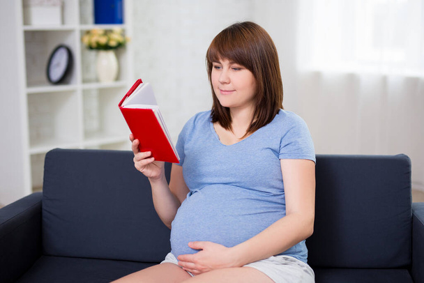 happy pregnant woman reading book in living room - Foto, Imagen